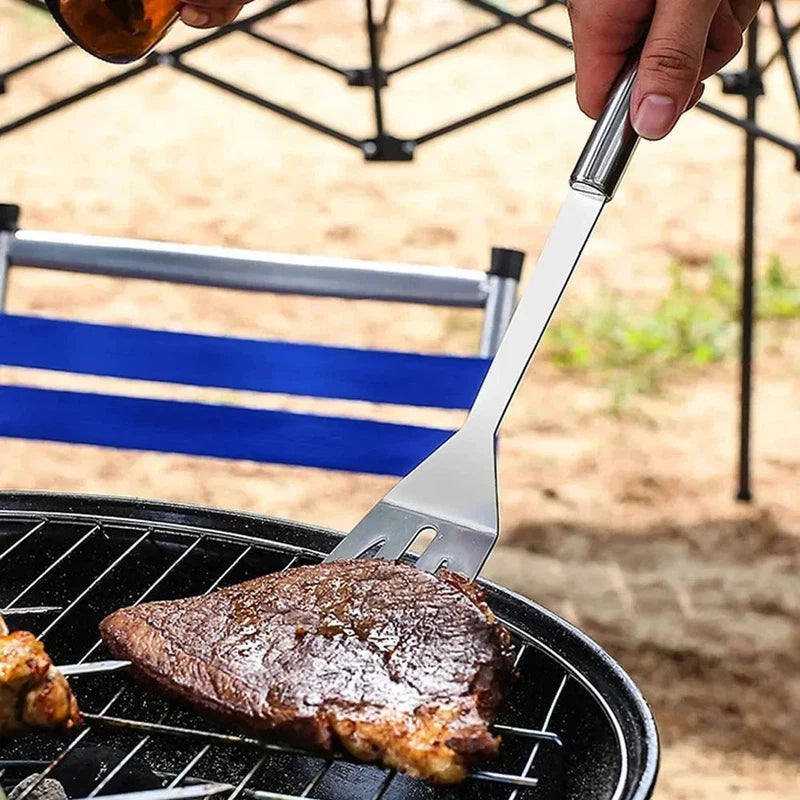 Stainless steel barbecue tools set for outdoor grilling, featuring a spatula in use over a grill with steak.